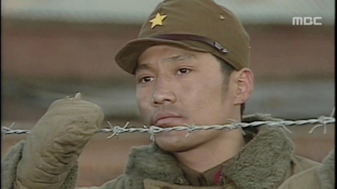 Choi Jae Seong como Choi Dae Chi Homem coreano com roupa militar de inverno durante a segunda guerra mundial, olha para fora de uma cerca de metal