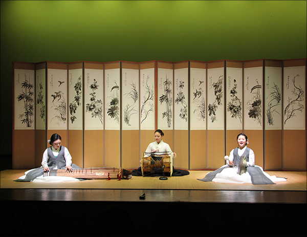 3 mulheres performando Seodo Sori. 3 mulheres sentadas e cada uma delas está tocando instrumentos coreanos antigos. Usam roupas tradicionais coreanas, conhecidas como hanbok. E estão tocando os instrumento gayageum e janggu. As 3 mulheres estão em um palco onde possui um cenário tradicional com telas de divisão de bambu com desenhos antigos, conhecidos como manhwa.