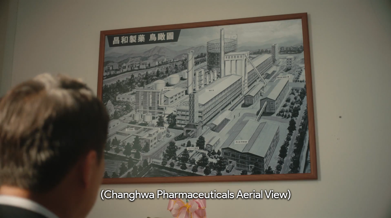 Foto onde um dos personagens olha para um quadro de uma fábrica. Com a legenda: "Vista Aérea para Farmacêutica Changhwa"