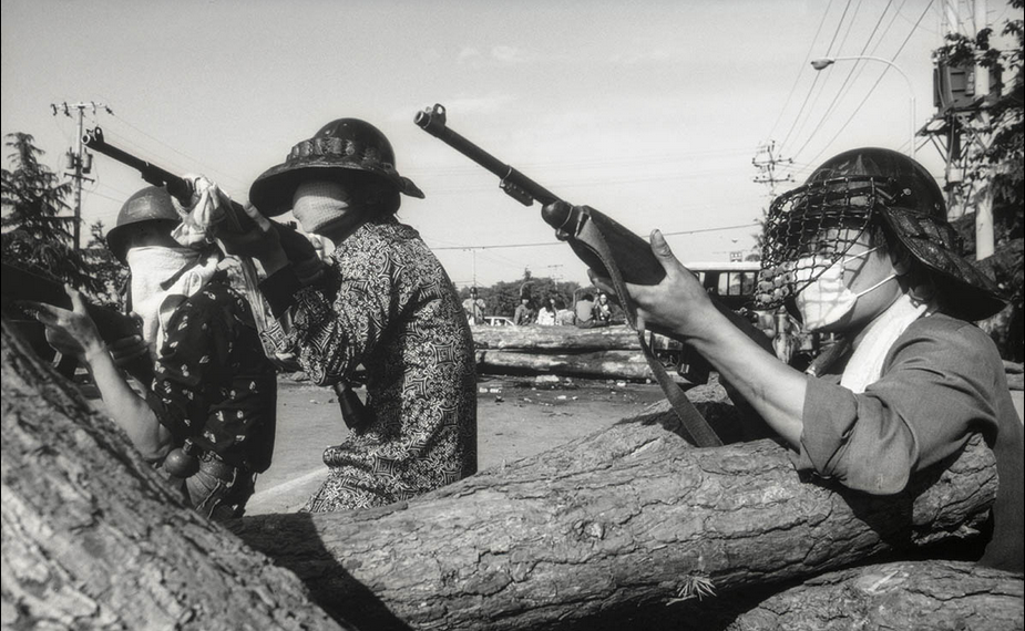 Foto de 3 pessoas com roupas casuais, capacetes de proteção, armas apontadas, mascaras brancas no rosto, atrás de troncos grandes de árvores.