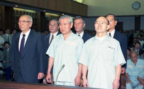 Advogados ao redor dos dois militares usando ternos pretos. Roh Tae Woo e Chun Doo Hwan com uniforme de penitenciária de cor azul claro.